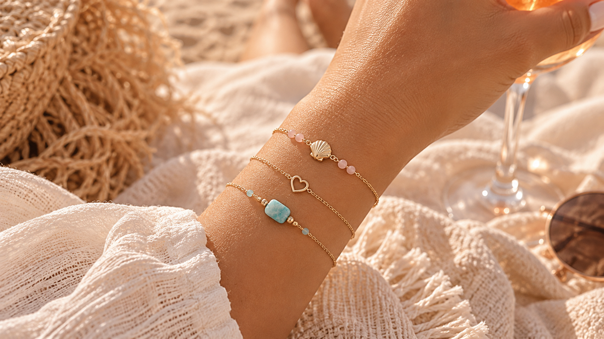 Trois bracelets dorés portés au poignet : coquillage avec pierres de soleil, petit cœur doré et bracelet avec pierre d'amazonite.